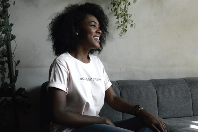 A smiling woman in a white t-shirt sitting on a grey couch.