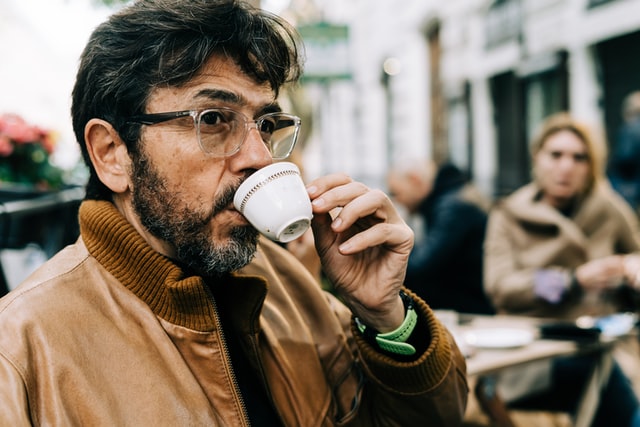 A man with grey hair and glasses sips coffee outside at a cafe.