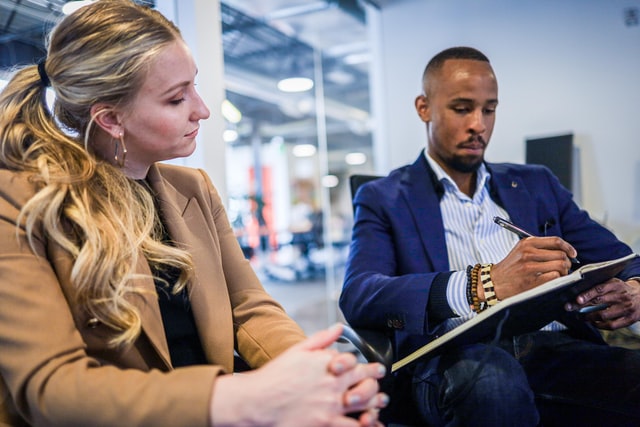 A woman watches as a man sitting next to her points to a notebook with a pen.