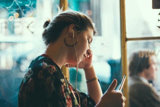 A girl wearing earbuds looks at a phone near a bright window.