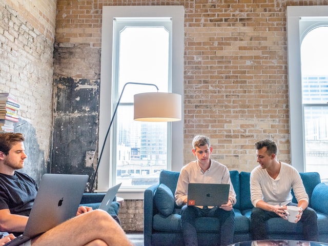 Three men sit on couches with laptops having a discussion.