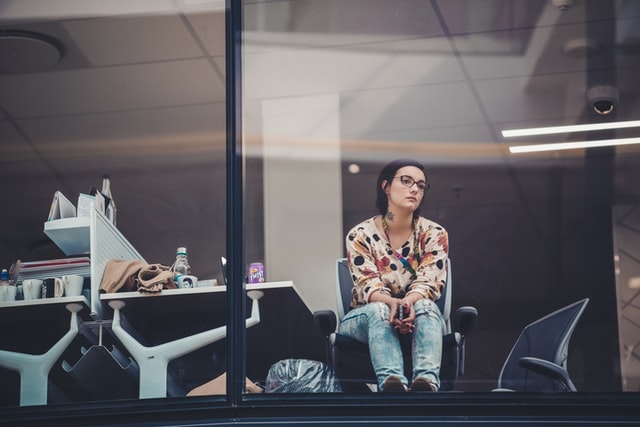 A dark-haired woman stares out the window from her desk at the office.