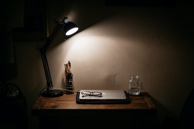 A computer, lamp, and a pair of glasses are on a desk at night.