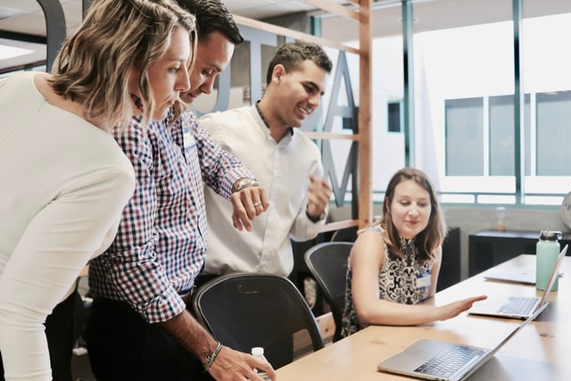 A team sits around a table, clapping for the woman who is standing.