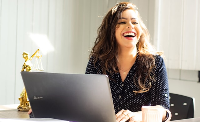 A woman laughs at her desk as she works on a laptop. 