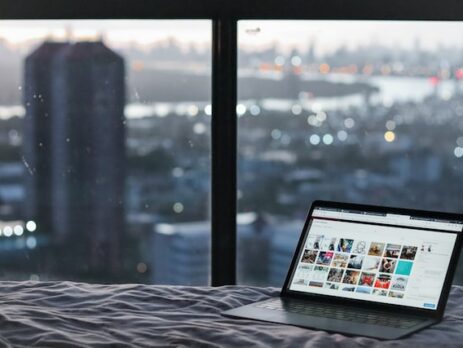 A computer sits on a bed near a large window. 