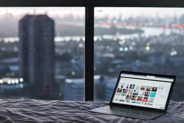 A computer sits on a bed near a large window. 