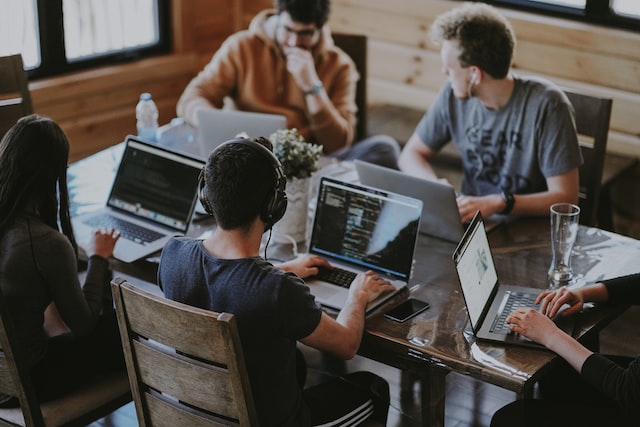 Five young individuals work on laptops around a table.