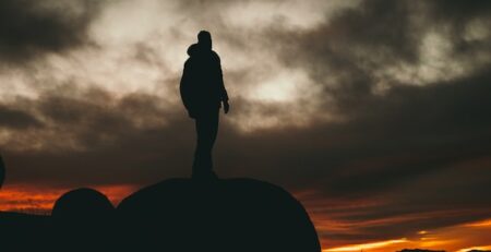 A man stands on a rock and looks out over the landscape in fading light. 