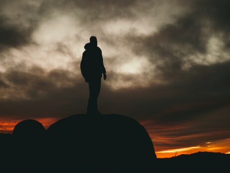 A man stands on a rock and looks out over the landscape in fading light. 