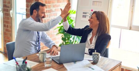 A man and a woman high-five as they sit at a table and work on laptops.