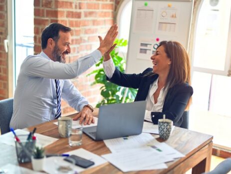 A man and a woman high-five as they sit at a table and work on laptops.