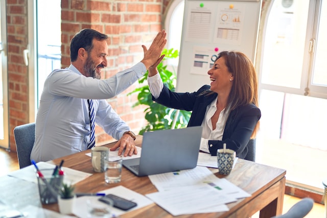 A man and a woman high-five as they sit at a table and work on laptops.