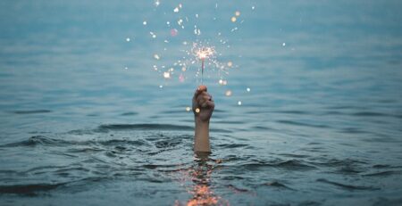 Alt-Text: A hand reaches out of water holding a lit sparkler. 