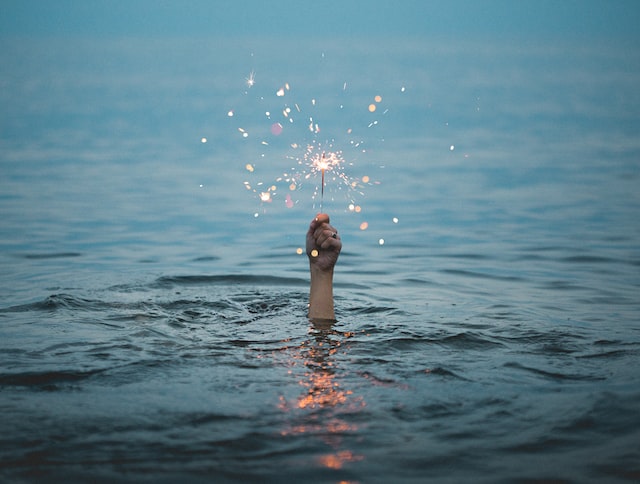 Alt-Text: A hand reaches out of water holding a lit sparkler. 
