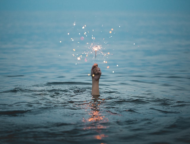 Alt-Text: A hand reaches out of water holding a lit sparkler. 