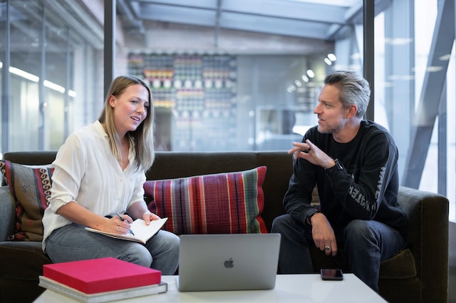 A blonde woman and gray haired man sit on a couch having a discussion.