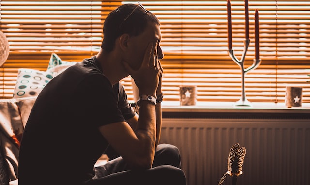 A man sits on a couch in a dim room with his head in his hands.