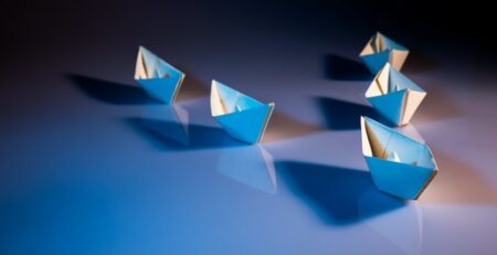 Five paper boats are on a table being led by a single paper boat.