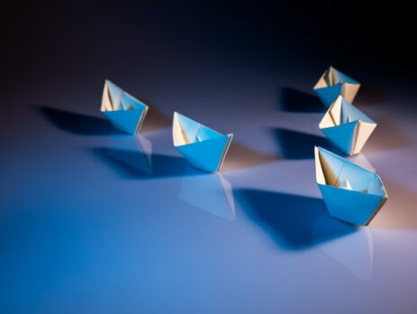 Five paper boats are on a table being led by a single paper boat.