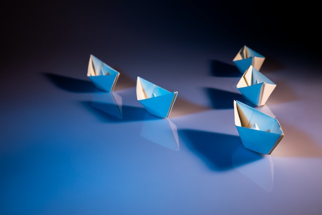 Five paper boats are on a table being led by a single paper boat.