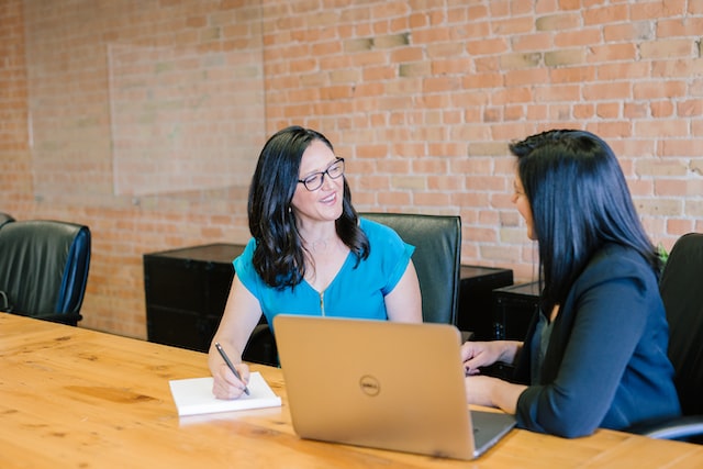 Two women discuss at a conference room table in front of a laptop.