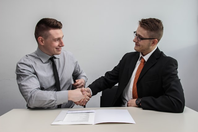 Two young men shake hands at a desk with a laptop on it.