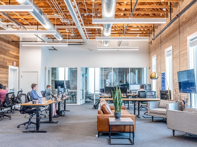 A view of an office room with empty desks as well as desks with people at them.