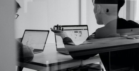 Two men discuss while seated at a table looking at laptops. 