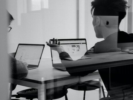 Two men discuss while seated at a table looking at laptops. 