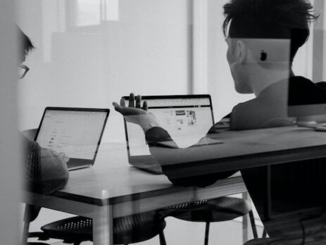 Two men discuss while seated at a table looking at laptops. 