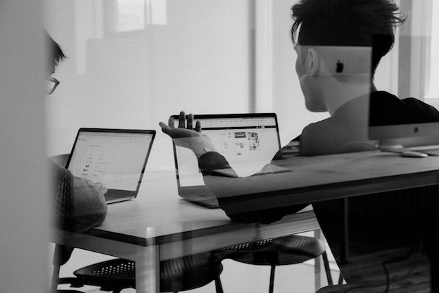 Two men discuss while seated at a table looking at laptops. 