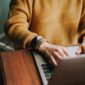 A person in an orange sweater works on a laptop.