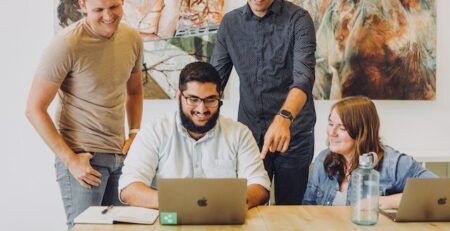 A manager stands behind an employee, recognizing him for a job well done while other employees are around.