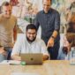 A manager stands behind an employee, recognizing him for a job well done while other employees are around.