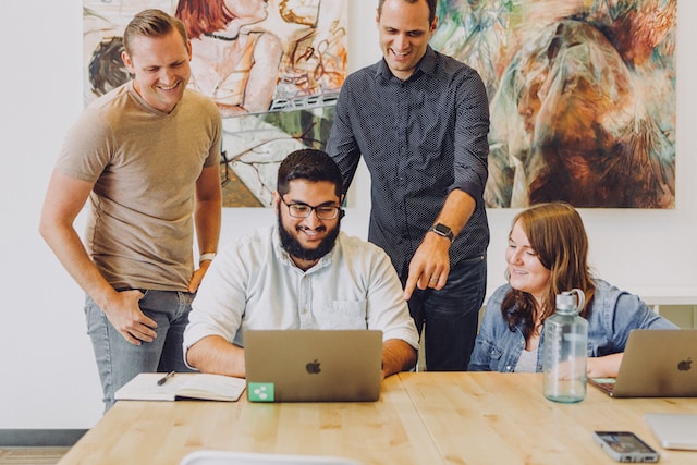 A manager stands behind an employee, recognizing him for a job well done while other employees are around.