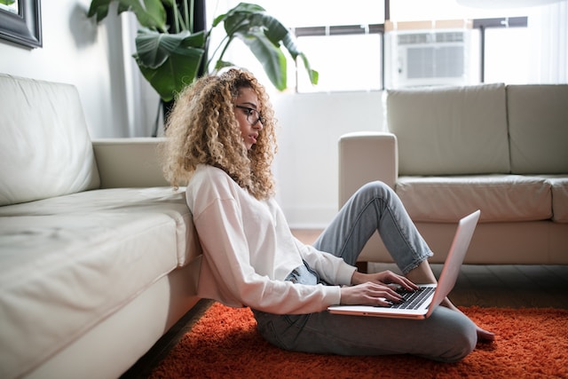 A woman sits on the floor with her back to the couch and a laptop on her lap.