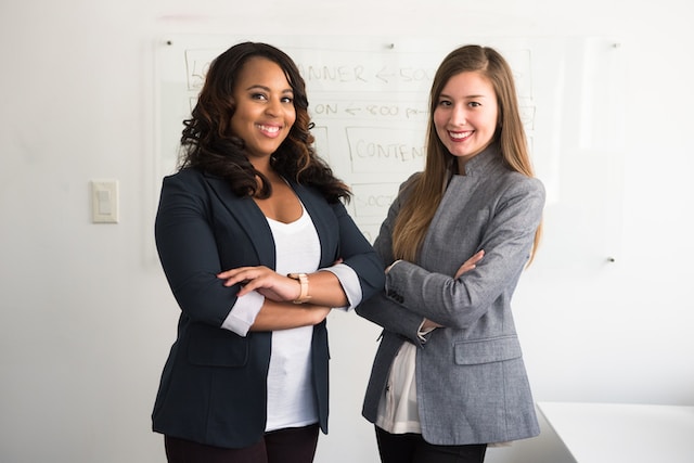 Two women in suits pose in front of a whiteboard. 