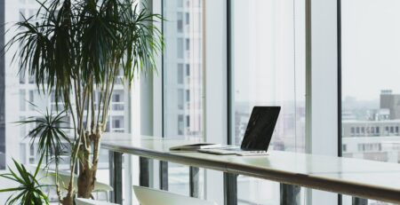 A laptop sits on a desk near the window in an empty workspace.