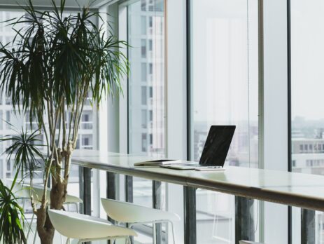 A laptop sits on a desk near the window in an empty workspace.