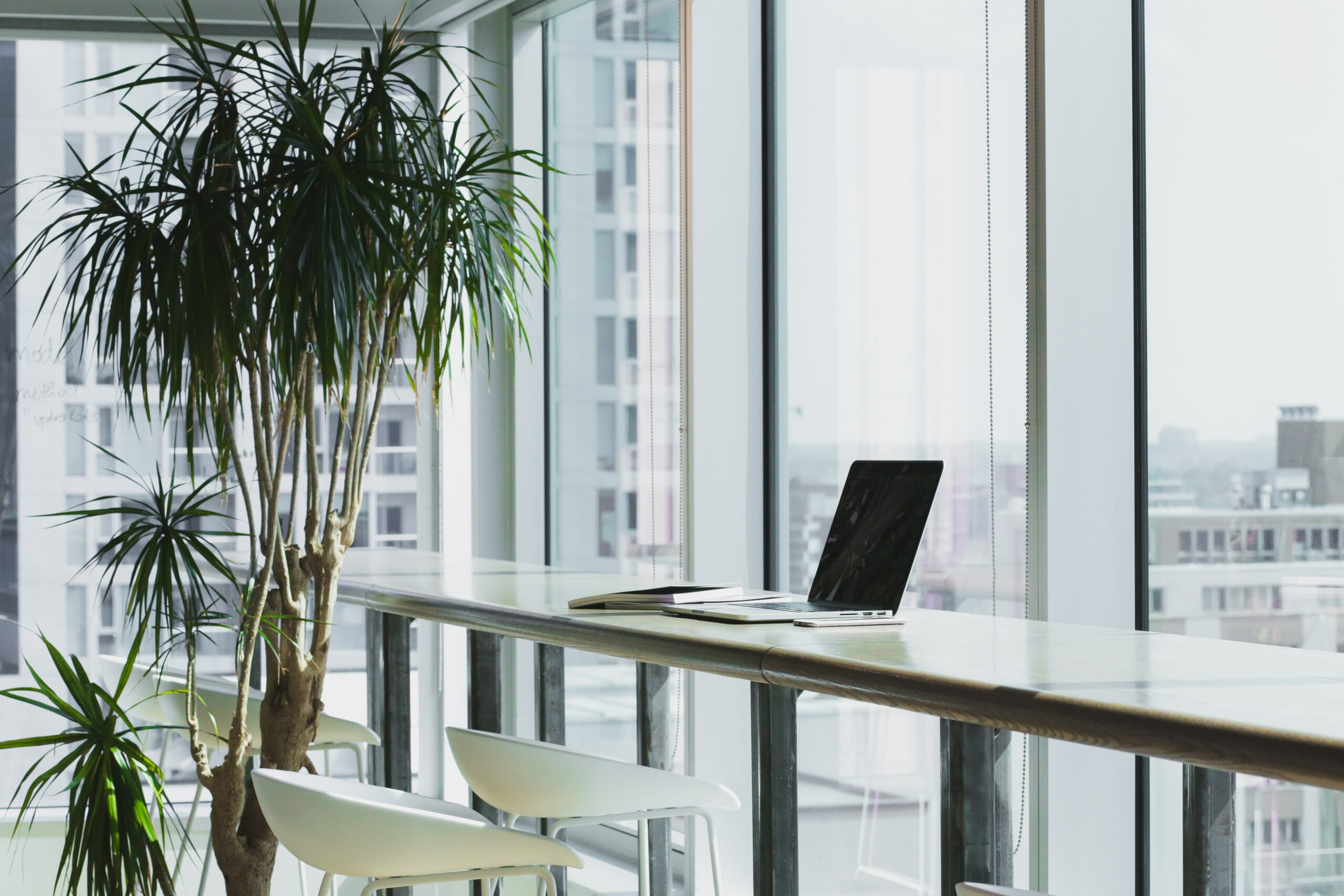 A laptop sits on a desk near the window in an empty workspace.