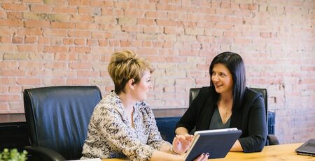 Two women sit at a table having a discussion while looking at a tablet. 