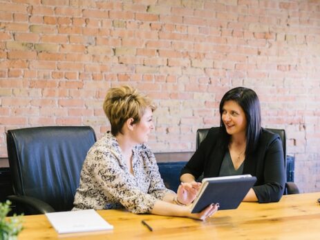 Two women sit at a table having a discussion while looking at a tablet. 