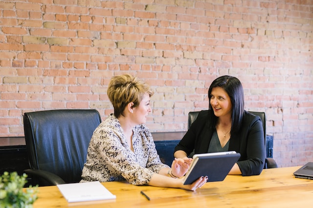 Two women sit at a table having a discussion while looking at a tablet. 