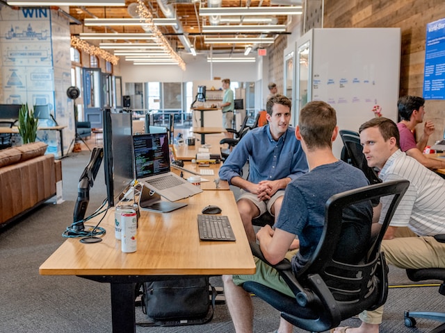 Three men surround a desk with a computer having a conversation.