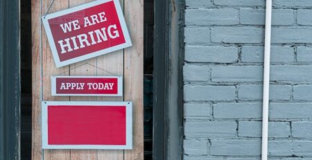 A red hiring sign is hung on a shop window above and apply today sign.