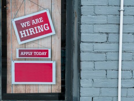 A red hiring sign is hung on a shop window above and apply today sign.
