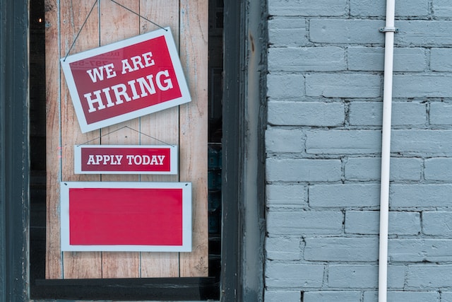 A red hiring sign is hung on a shop window above and apply today sign.