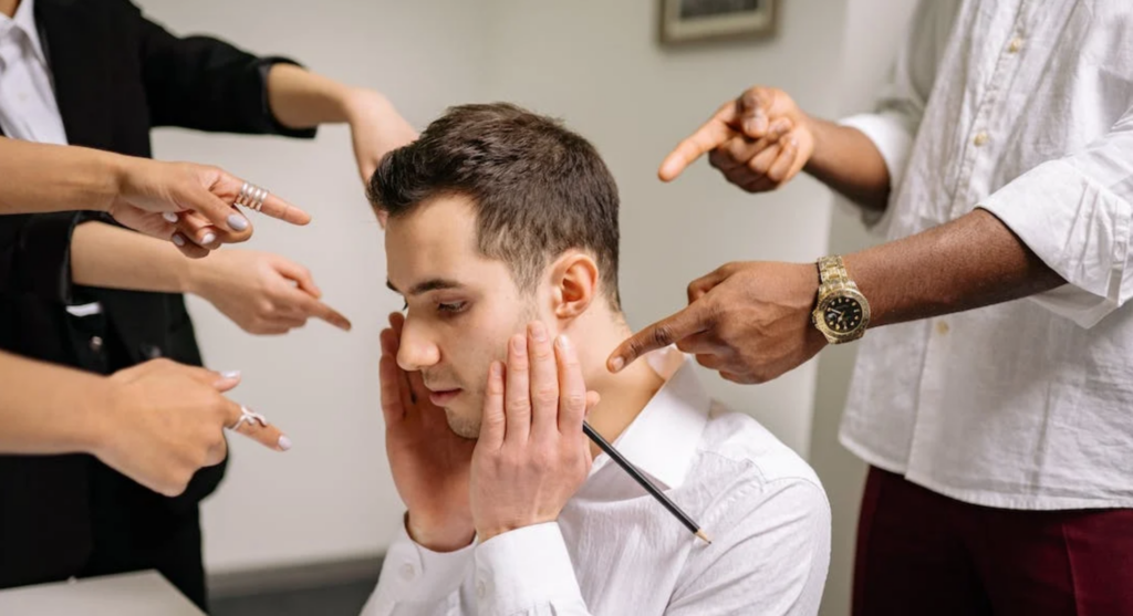 Man sitting and holding his face while colleagues point their fingers at him.