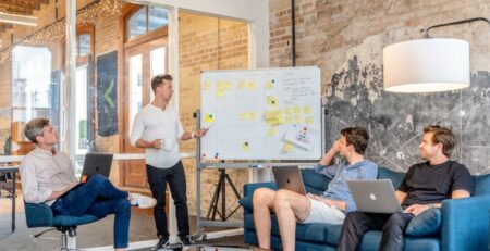 Three men using their laptops in a meeting while watching a man use a whiteboard.